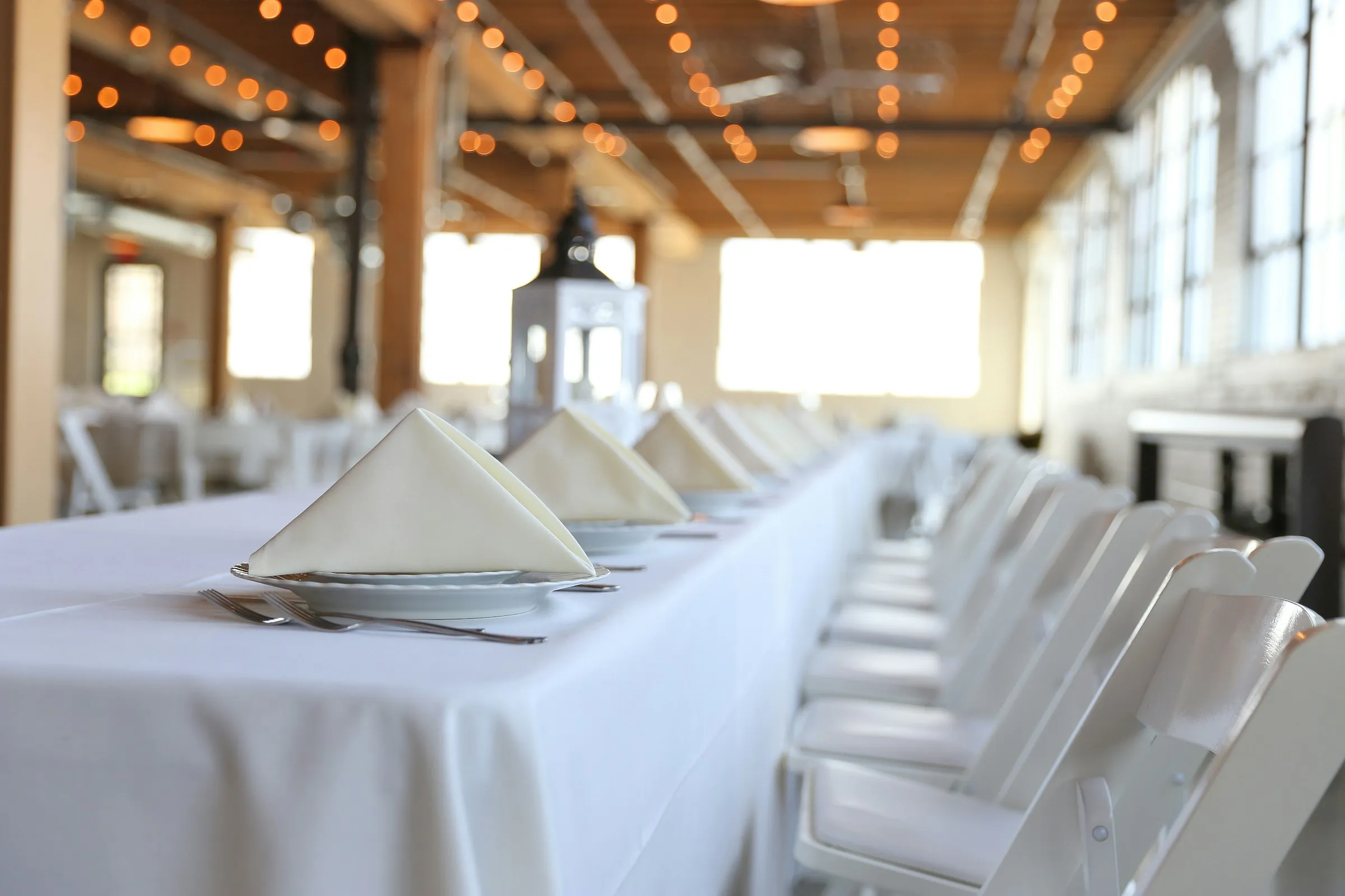 A long table set with white linens and folded napkins under warm string lights